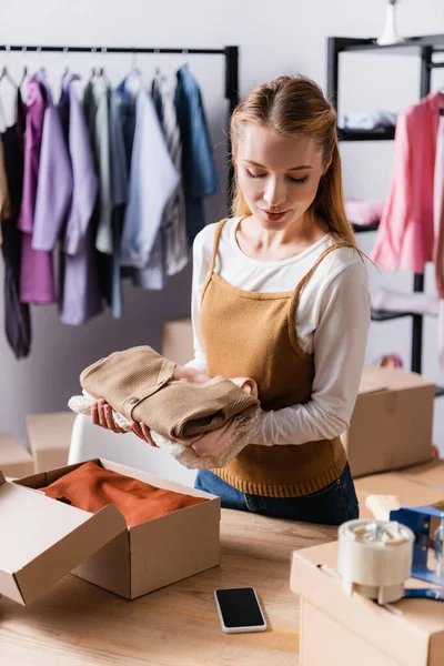 Showroom owner holding clothes near cardboard boxes and smartphone, blurred foreground — Stock Photo