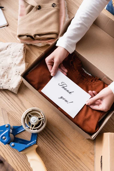 Partial view of seller holding card with thank you lettering near box with clothes in showroom — Stock Photo