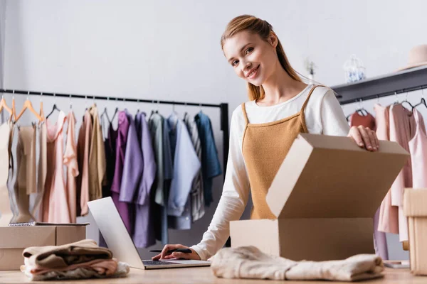 Propriétaire réussi showroom souriant à la caméra près de boîtes en carton et cintres avec des vêtements sur fond flou — Photo de stock