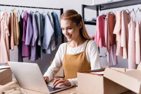 Proprietario showroom sorridente digitando sul computer portatile vicino scatole di cartone e rack su sfondo sfocato — Foto stock