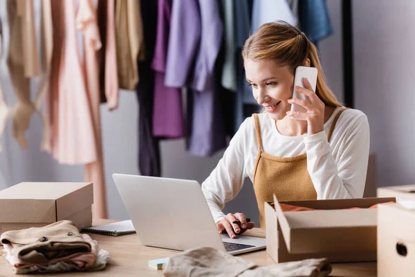 Positive showroom owner accepting order on smartphone near laptop, blurred foreground — Stock Photo