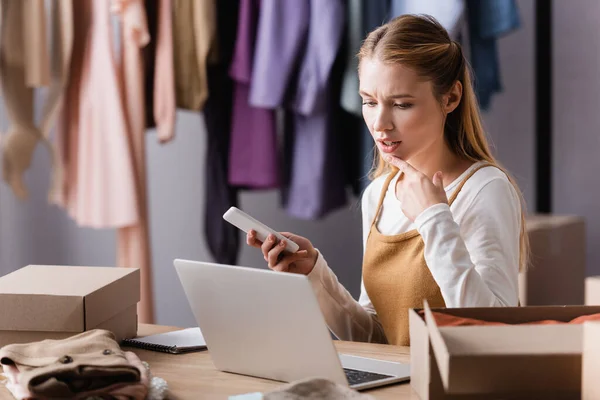 Vendeur sérieux regardant ordinateur portable près des boîtes en carton dans la boutique de mode, au premier plan flou — Photo de stock