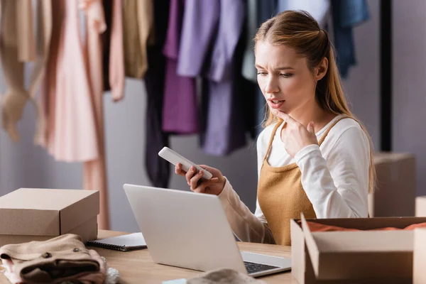 Serious seller looking at laptop near carton boxes in fashion boutique, blurred foreground — Stock Photo