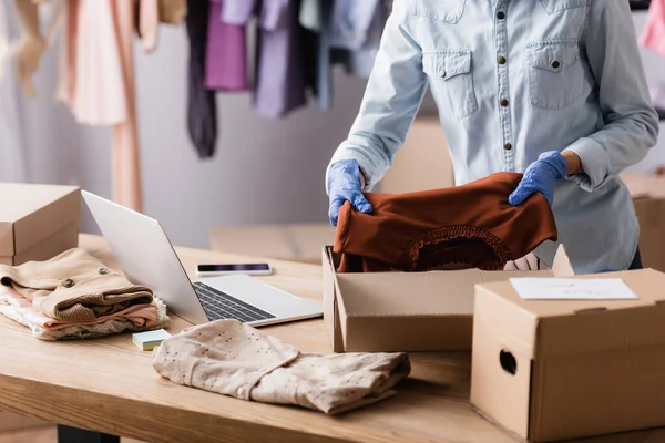 Cropped view of seller in latex gloves packing clothes into boxes in fashion boutique — Stock Photo