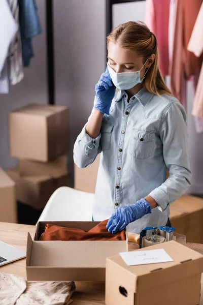 Showroom proprietor in safety mask, talking on smartphone while working with packages on blurred foreground — Stock Photo