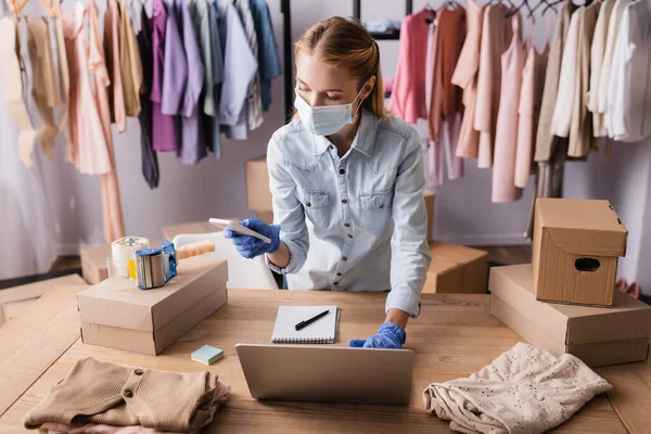 Seller in medical mask, working with laptop and mobile phone near cardboard boxes in showroom — Stock Photo