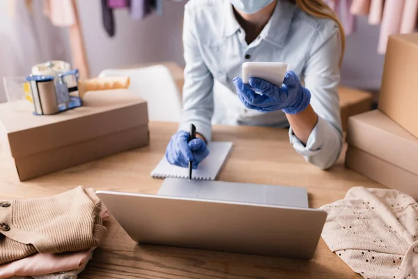 Partial view of proprietor in latex gloves working with smartphone and laptop in showroom, blurred background — Stock Photo