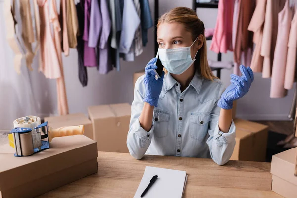 Young seller in medical mask, talking on smartphone at workplace in showroom — Stock Photo