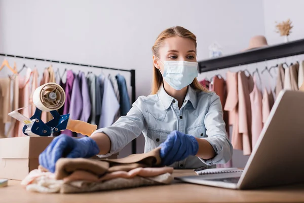 Vendedor joven en máscara médica, trabajando con ropa cerca de la computadora portátil en la sala de exposición en primer plano borrosa - foto de stock