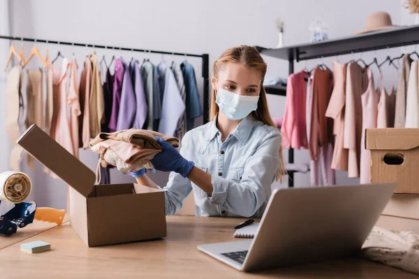 Proprietário boutique de moda em máscara médica, embalagem de roupas em caixas perto de laptop em primeiro plano borrado — Fotografia de Stock