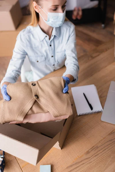 Vista de ángulo alto del propietario de la sala de exposición en máscara médica, sosteniendo la ropa cerca de la caja sobre fondo borroso - foto de stock