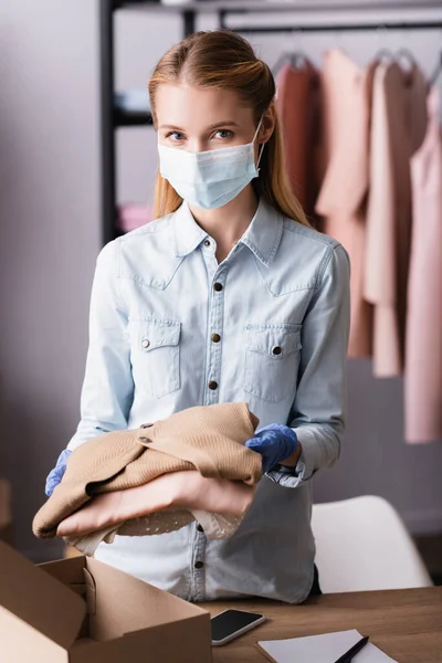 Young proprietor in medical mask, holding clothes and looking at camera in showroom — Stock Photo