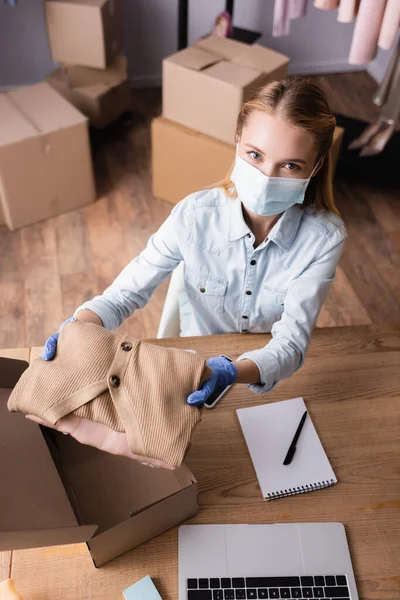 High angle view of seller in medical mask, holding clothes and looking at camera in showroom — Stock Photo