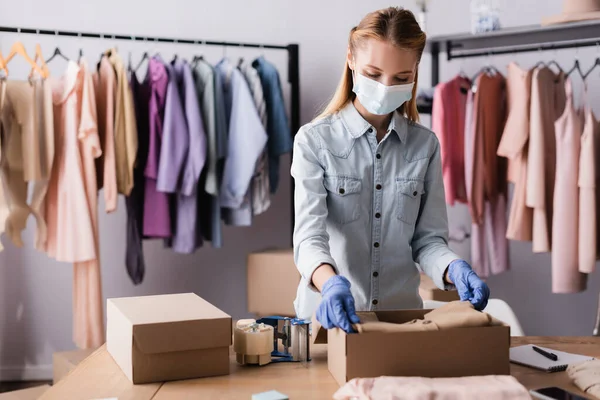 Jovem empresária em máscara médica, embalando roupas em caixas no showroom, foreground borrado — Fotografia de Stock