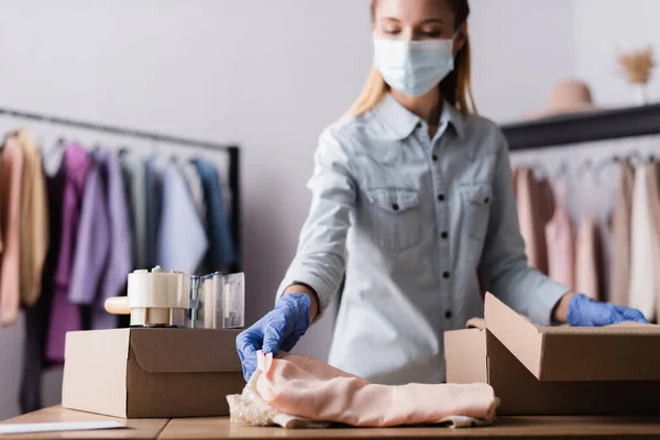 Owner of fashion boutique in medical mask, packing clothes into box on blurred background — Stock Photo