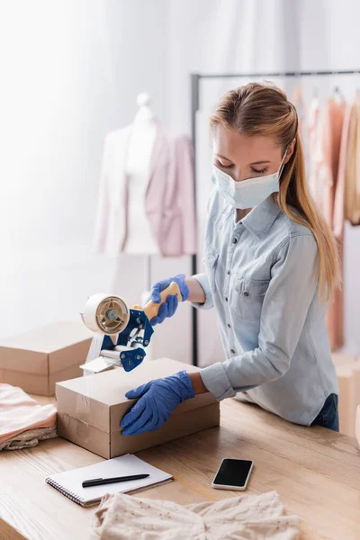 Showroom proprietor in protective mask, holding adhesive tape near box and smartphone with blank screen — Stock Photo