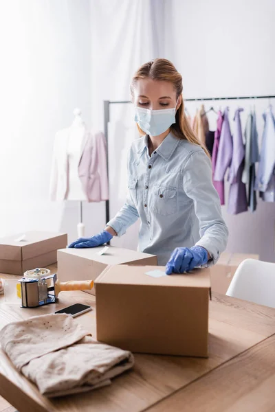 Young proprietor in medical mask, fixing sticky notes on boxes in showroom — Stock Photo