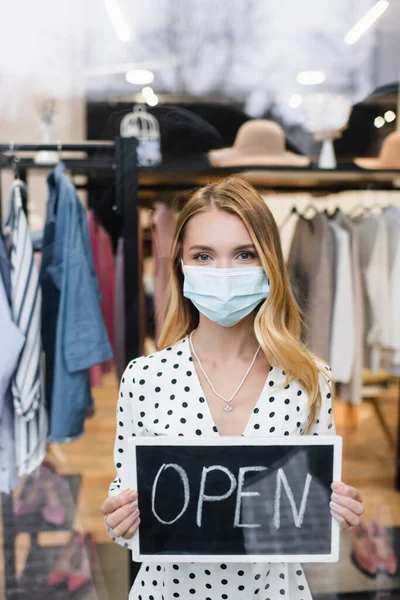 Positive businesswoman in medical mask, holding board with open lettering in fashion boutique — Stock Photo
