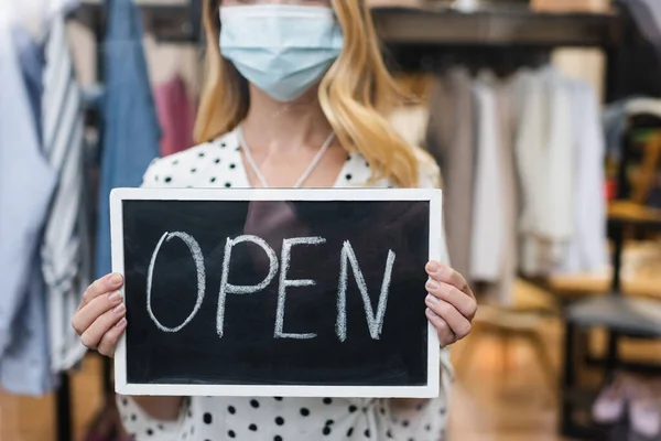 Vista parcial do proprietário do showroom na máscara médica, segurando a placa com letras abertas, fundo borrado — Fotografia de Stock