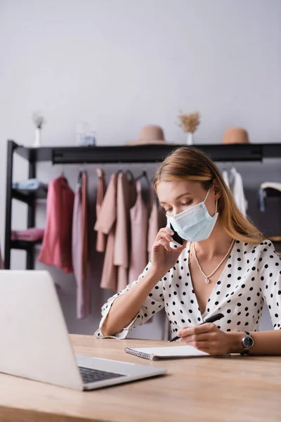 Mujer de negocios en máscara médica, hablando en el teléfono inteligente cerca de la computadora portátil en la sala de exposición - foto de stock