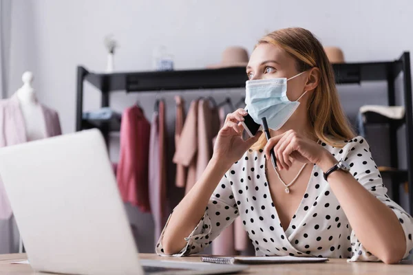 Propietario de la sala de exposición en máscara médica, hablando en el teléfono inteligente cerca de la computadora portátil en primer plano borrosa — Stock Photo
