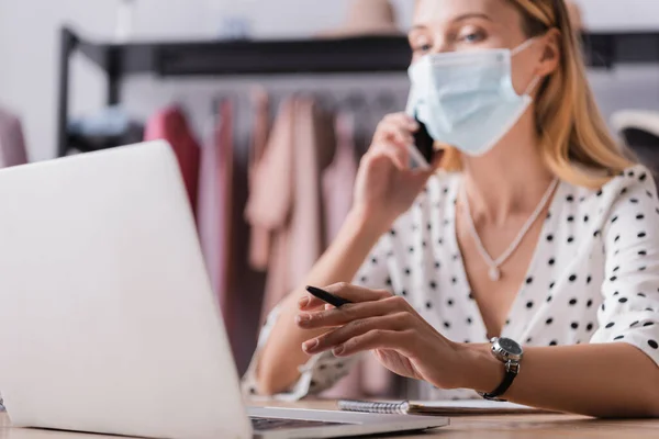 Бизнесвумен в медицинской маске принимает заказ в выставочном зале, работая рядом с ноутбуком на размытом фоне — стоковое фото