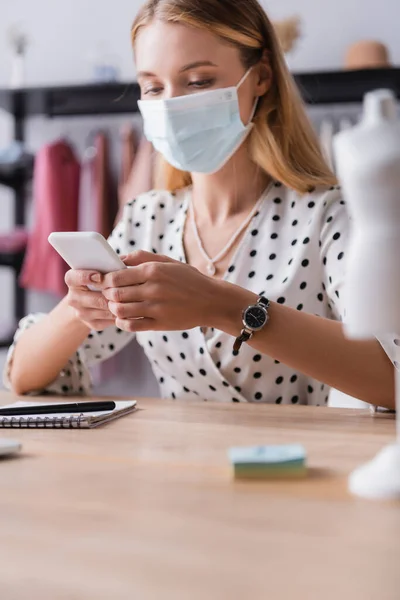 Propriétaire du showroom en masque de protection, messagerie sur smartphone sur le lieu de travail au premier plan flou — Photo de stock