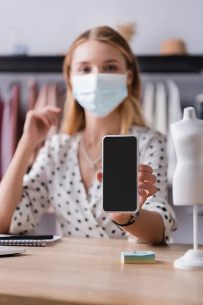 Propietario de la sala de exposición en máscara médica, mostrando teléfono inteligente con pantalla en blanco sobre fondo borroso - foto de stock