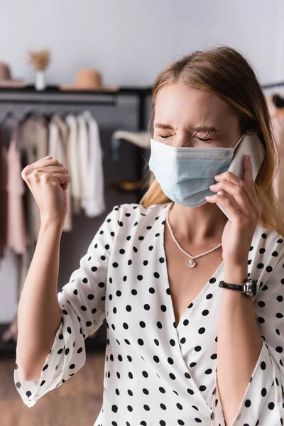 Propietario enojado en máscara médica sosteniendo el puño apretado mientras habla en el teléfono móvil en la sala de exposición - foto de stock