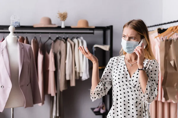 Upset showroom proprietor in medical mask talking on mobile phone near mannequin and hangers on blurred background — Stock Photo