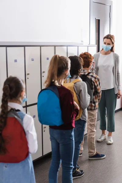 Teacher holding non contact thermometer near pupils in medical masks on blurred foreground in corridor — Stock Photo
