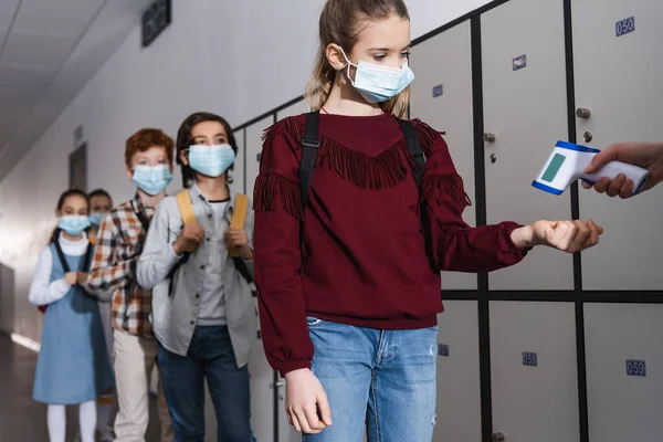 Chica en máscara médica que mide la temperatura cerca del profesor con termómetro sin contacto y compañeros de clase en la escuela - foto de stock