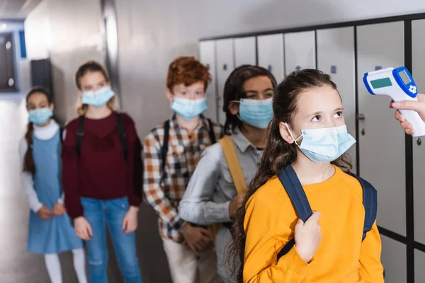 Profesor usando termómetro infrarrojo en la pupila en máscara médica cerca de niños con fondo borroso - foto de stock