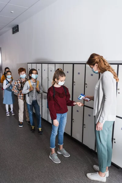 Teacher in medical mask checking temperature of pupil with infrared thermometer in corridor — Stock Photo