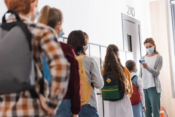 Teacher in medical mask using infrared thermometer on schoolchild with backpack in hall — Stock Photo