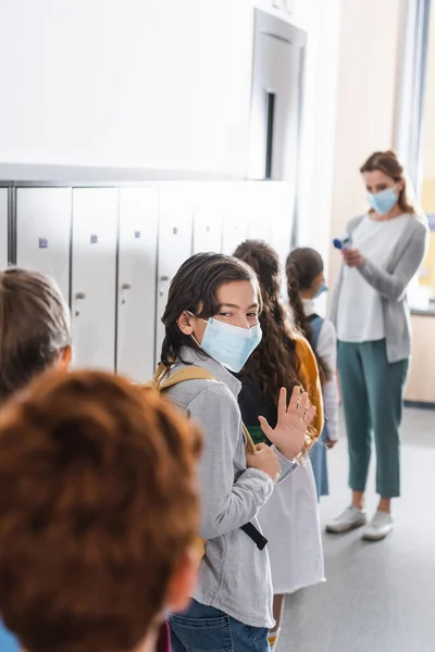 Boy in medical mask waving hand near classmates and teacher with infrared thermometer on blurred background — Stock Photo