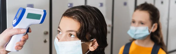 Insegnante che misura la temperatura della pupilla in maschera medica vicino a scolara su sfondo sfocato, banner — Foto stock