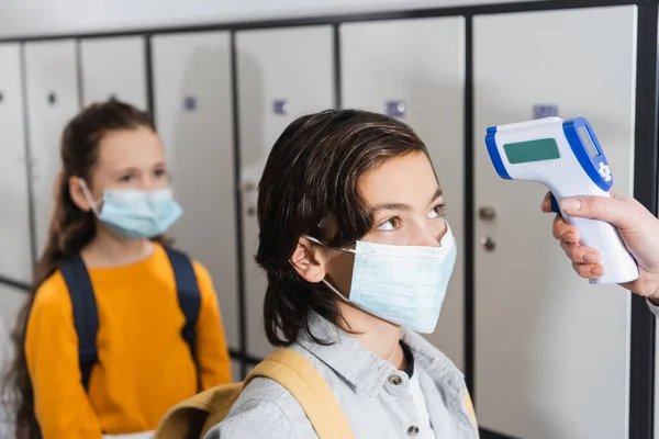 Profesor con termómetro infrarrojo comprobando la temperatura del niño en máscara protectora en la sala - foto de stock