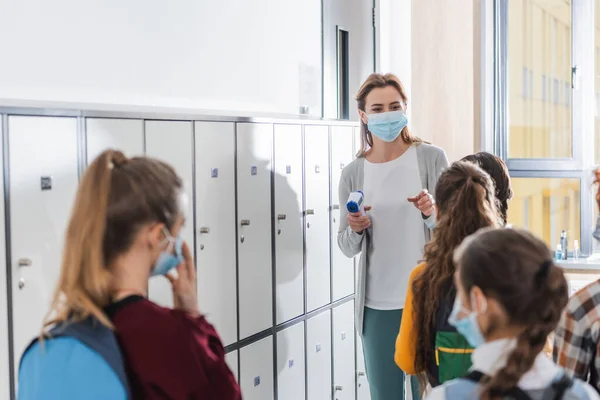 Teacher with infrared thermometer pointing with finger at kids in medical masks on blurred foreground in school — Stock Photo