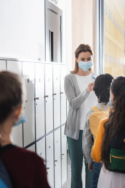 Profesor en máscara médica midiendo la temperatura de los alumnos cerca de casilleros en la escuela - foto de stock