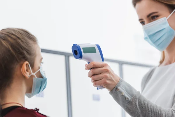 Berührungsloses Thermometer in der Hand des Lehrers in medizinischer Maske zur Temperaturmessung der Schülerin auf verschwommenem Vordergrund — Stockfoto