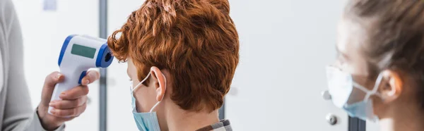 Schoolboy in medical mask standing near teacher with infrared thermometer and classmate on blurred foreground, banner — Stock Photo