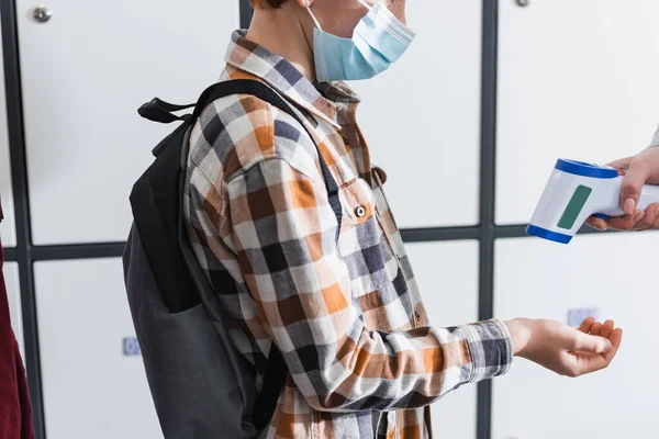 Ausgeschnittene Ansicht eines Schülers in medizinischer Maske, der in der Nähe des Lehrers steht und die Temperatur mit Infrarot-Thermometer misst, Banner — Stockfoto