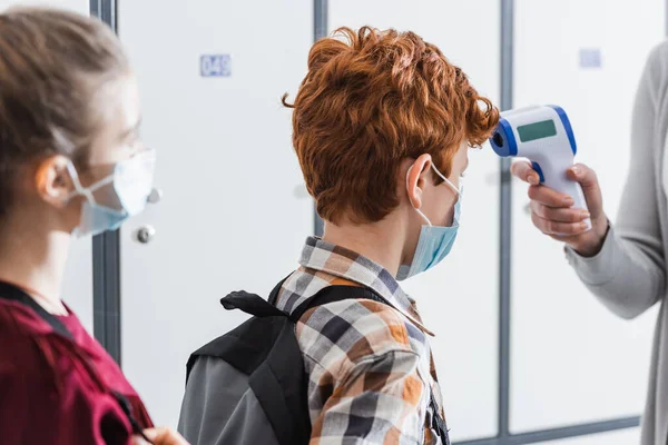 Teacher using infrared thermometer on schoolboy in protective mask with backpack — Stock Photo