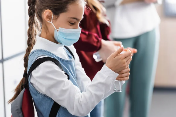Studentessa in maschera medica utilizzando disinfettante mano vicino compagno di classe e insegnante su sfondo sfocato — Foto stock