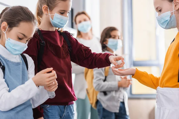 Школярі в медичних масках використовують дезінфікуючий засіб для рук біля вчителя на розмитому тлі — стокове фото