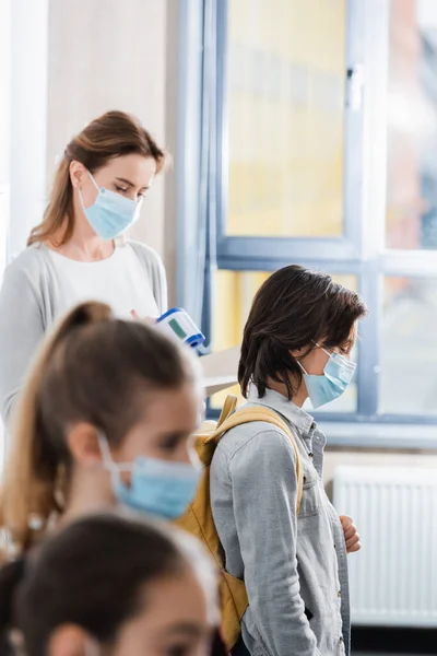 Colegial en máscara médica de pie cerca del profesor con termómetro infrarrojo y compañeros de clase - foto de stock