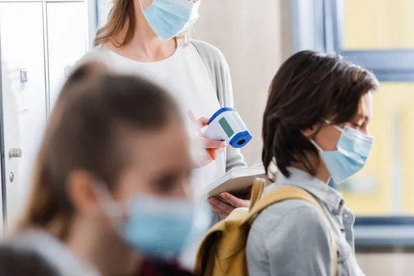 Profesor sosteniendo termómetro infrarrojo y cuaderno cerca de los alumnos con máscaras médicas en primer plano borroso - foto de stock