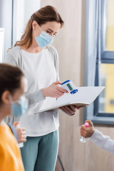 Teacher in medical mask holding infrared thermometer and notebook near pupils on blurred foreground — Stock Photo