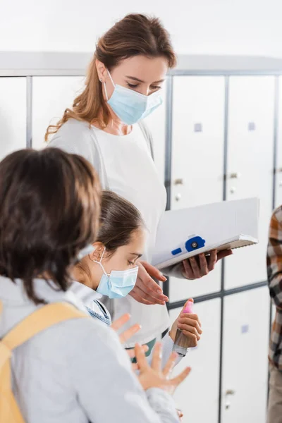 Teacher with notebook and infrared thermometer standing near pupils in protective masks with disinfector — Stock Photo